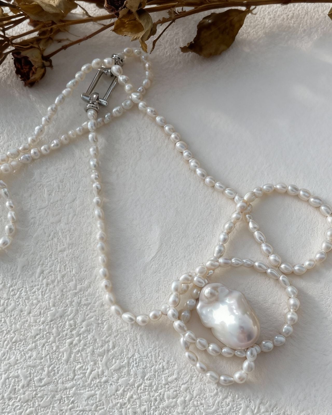 Pearl necklace with a decorative clasp on a textured white surface with dried flowers.