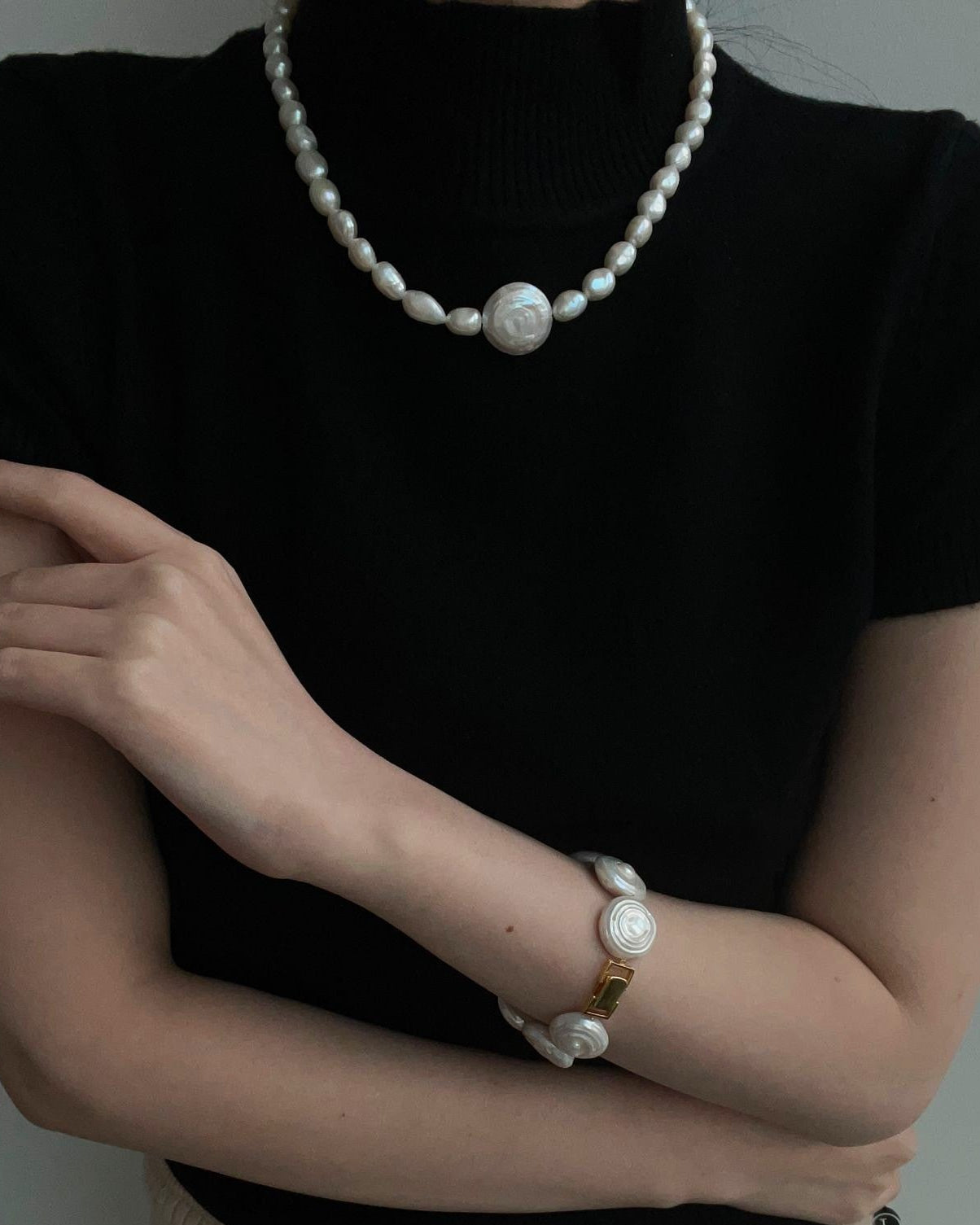 Person wearing a black top and beige pants with pearl jewelry on a neutral background