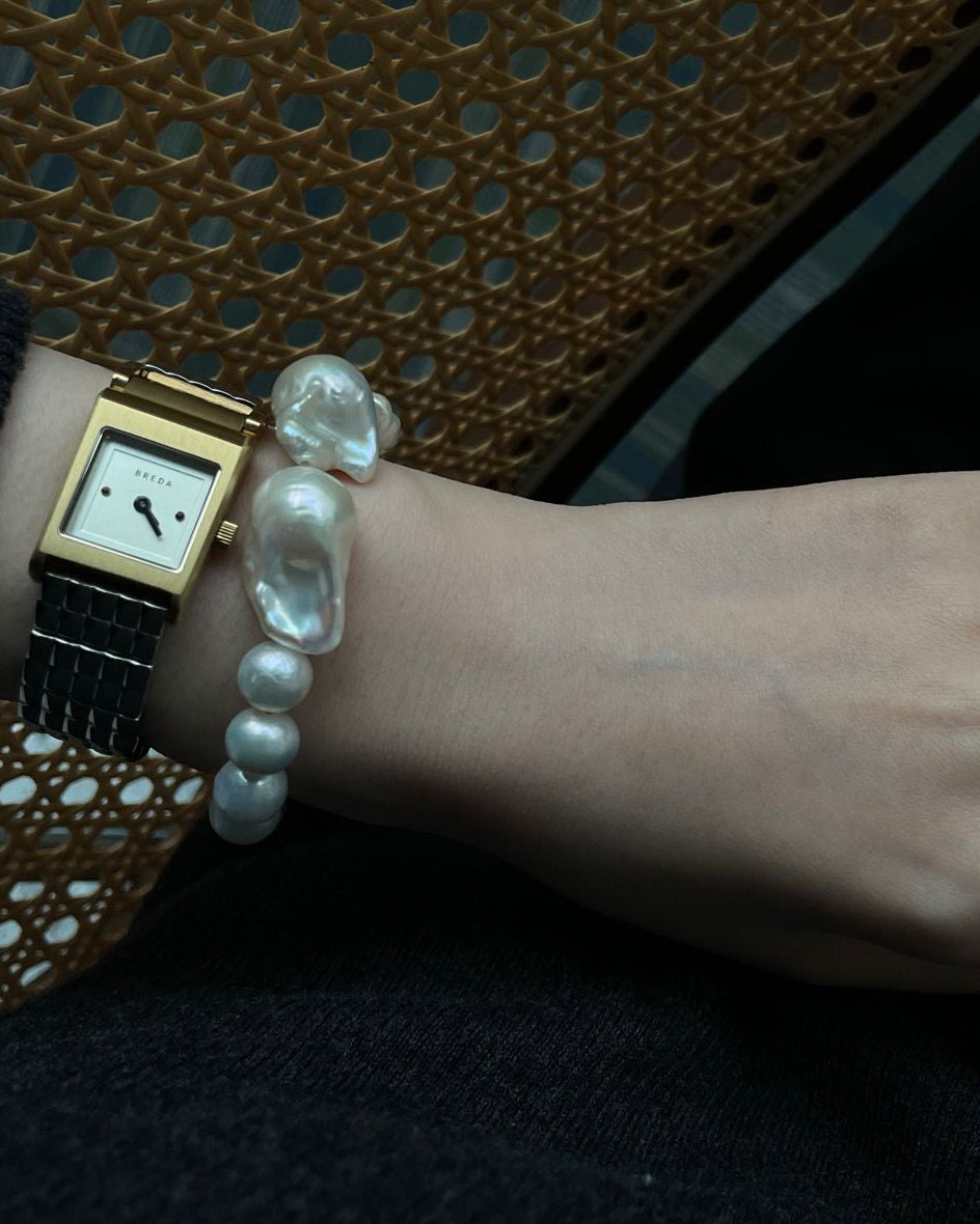 Hand wearing a pearl bracelet and ring with a gold watch, against a dark background.
