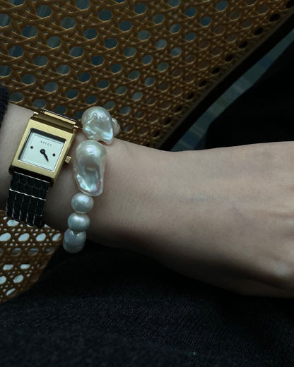 Hand wearing a pearl bracelet and ring with a gold watch, against a dark background.