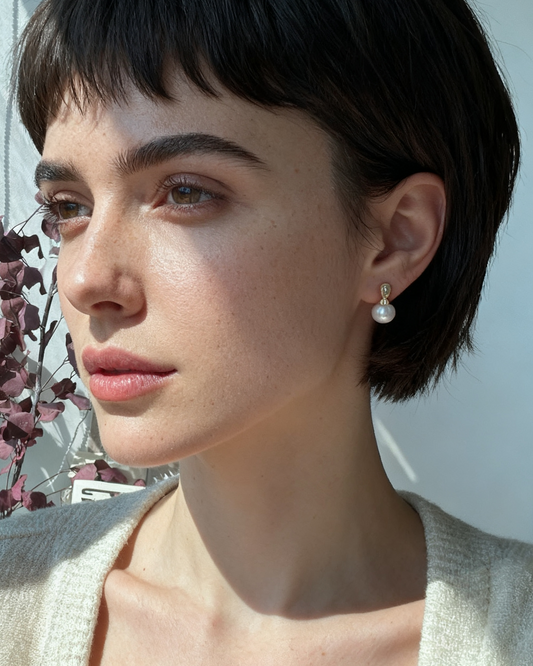 Woman with short dark hair wearing pearl earrings, standing near a window with flowers.